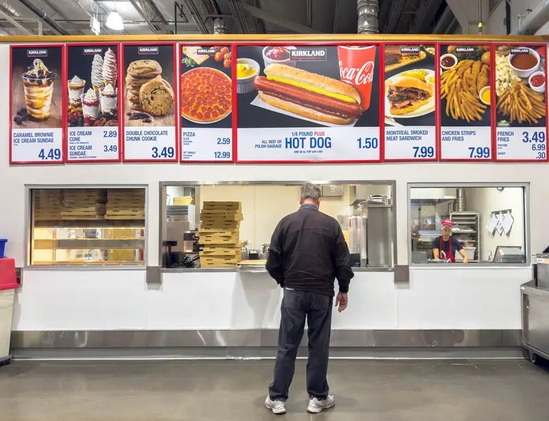 A customer waits at a Costco food court counter beneath a large menu board showing prices