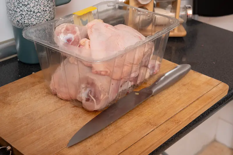 Raw chicken pieces in packaging with knife on wooden board